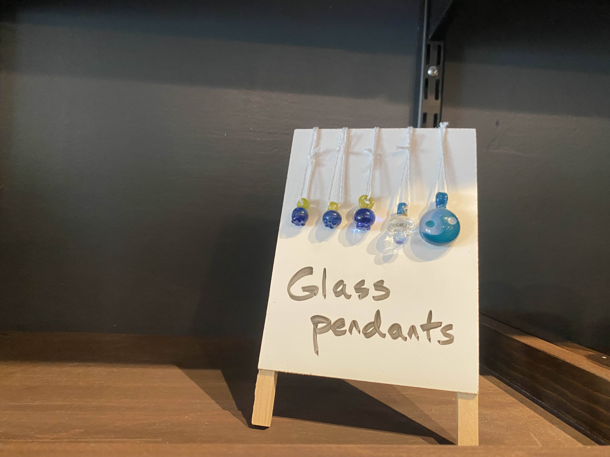 Zoomed in photo of a portion of the wooden merchandise shelf. Showcases 5 glass pendants hanging over a mini easel that has the words 'Glass pendants' written on it in black marker. Three of the pendants are blue and blueberry shaped, one is clear and mushroom shaped, and the last is light blue and depicts a yin yang symbol.
