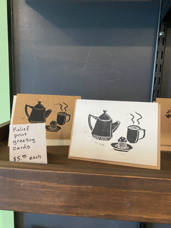 Zoomed in photo of a portion of the wooden merchandise shelf. Showcases greeting cards depicting a teapot, steaming hot cup of tea, and a muffin on a plate printed with black ink. To the left of the cards is signage marketing them as 'Relief print greeting cards' being sold for $5.00 each.