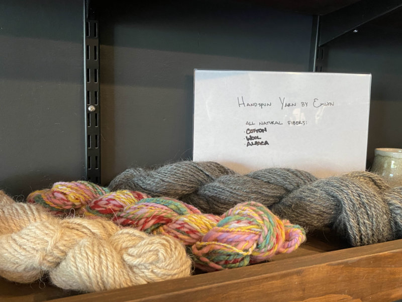 Zoomed in photo of a portion of the wooden merchandise shelf. Showcases various handspun yarns in different materials and colors.