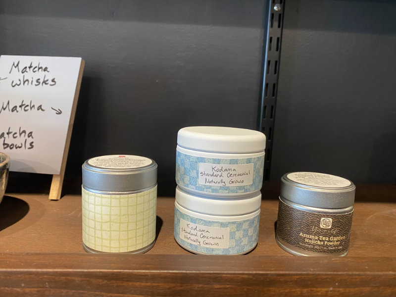 Zoomed in photo of a portion of the wooden merchandise shelf. Showcases 4 small tins labeled as matcha and hojicha powders.