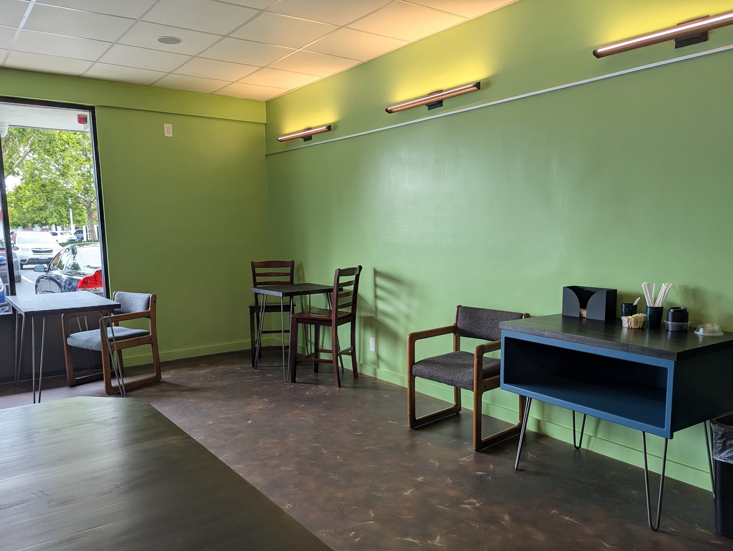 Photo of the Fair Grounds building interior, facing the shop front. Shows a window facing the parking lot as well as several wooden tables and chairs, and a self-service counter with straws, napkins, lids, and sugar.